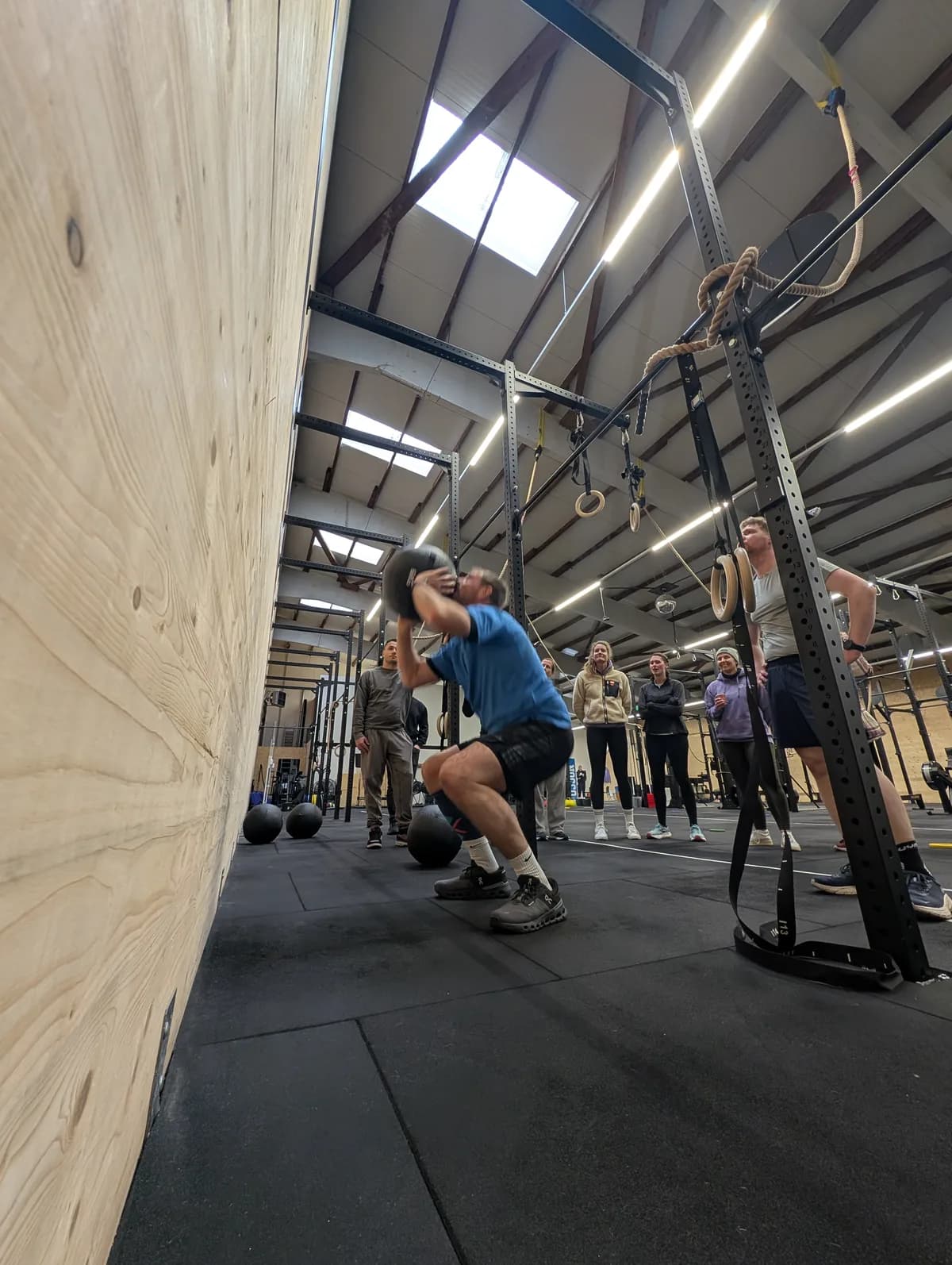 Athlet fängt den Wall Ball an der Holzwand, im Hintergrund verfolgt eine Gruppe Zuschauer den Lauf — HYROX Race Simulation bei AMITY Athletics